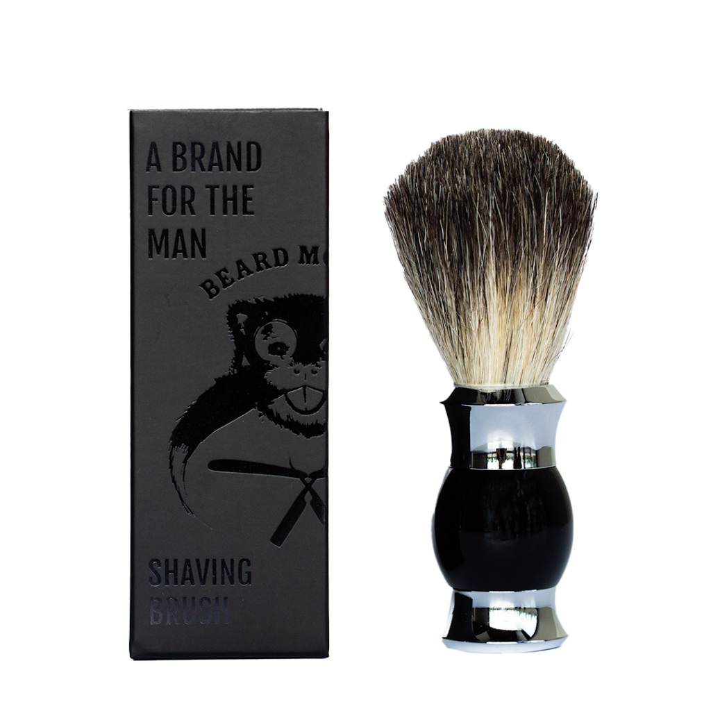 Rakborste från Beard Monkey med svart, blankt handtag i kombination med silverfärgade detaljer och tät borst i ljusa och mörka toner. Rakborsten visas bredvid en mörk Beard Monkey-förpackning med diskret grafisk design.