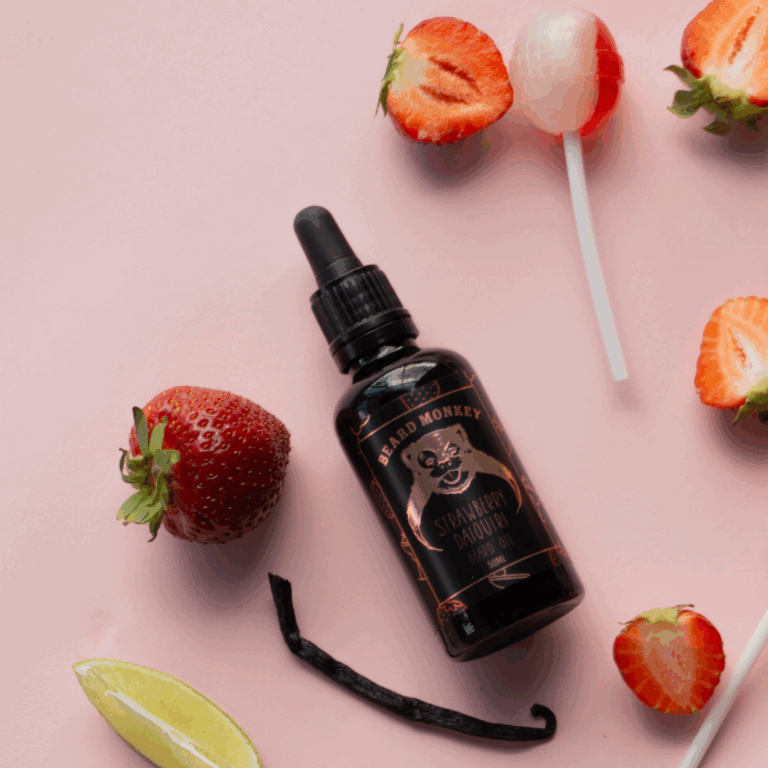 En skäggolja i pipettflaska märkt "Beard Monkey Strawberry Daiquiri" omgiven av jordgubbar, klubbor, en limeklyfta och en vaniljstång på en rosa bakgrund.