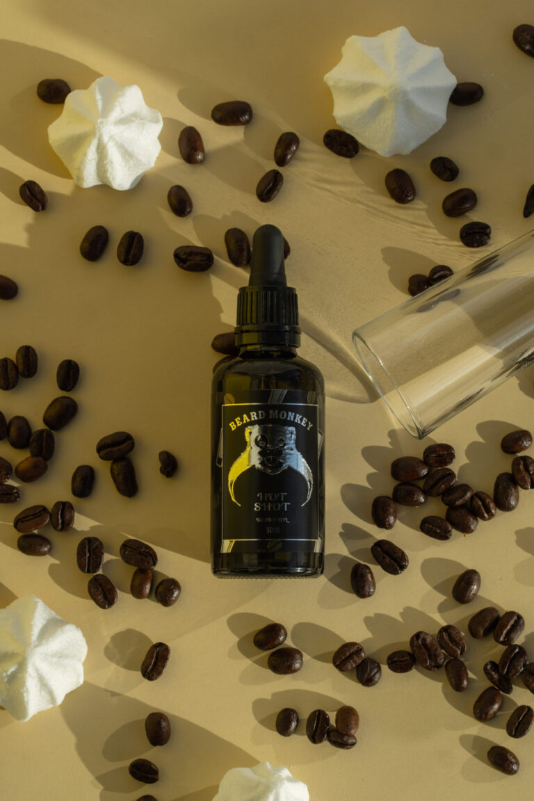 En flaska Beard Monkey skäggolja med pipett ligger på en beige yta, omgiven av kaffebönor, maränger och ett klart snapsglas.