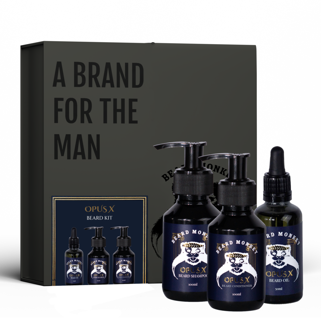 Mörk presentförpackning från Beard Monkey med svarta Beard Monkey-detaljer. Framför förpackningen står tre produkter i OPUS X-serien: ett skäggschampo och ett skäggbalsam i svarta pumpflaskor samt en skäggolja i svart pipettflaska, alla med etiketter med blå bakgrund och guldiga samt vita detaljer.